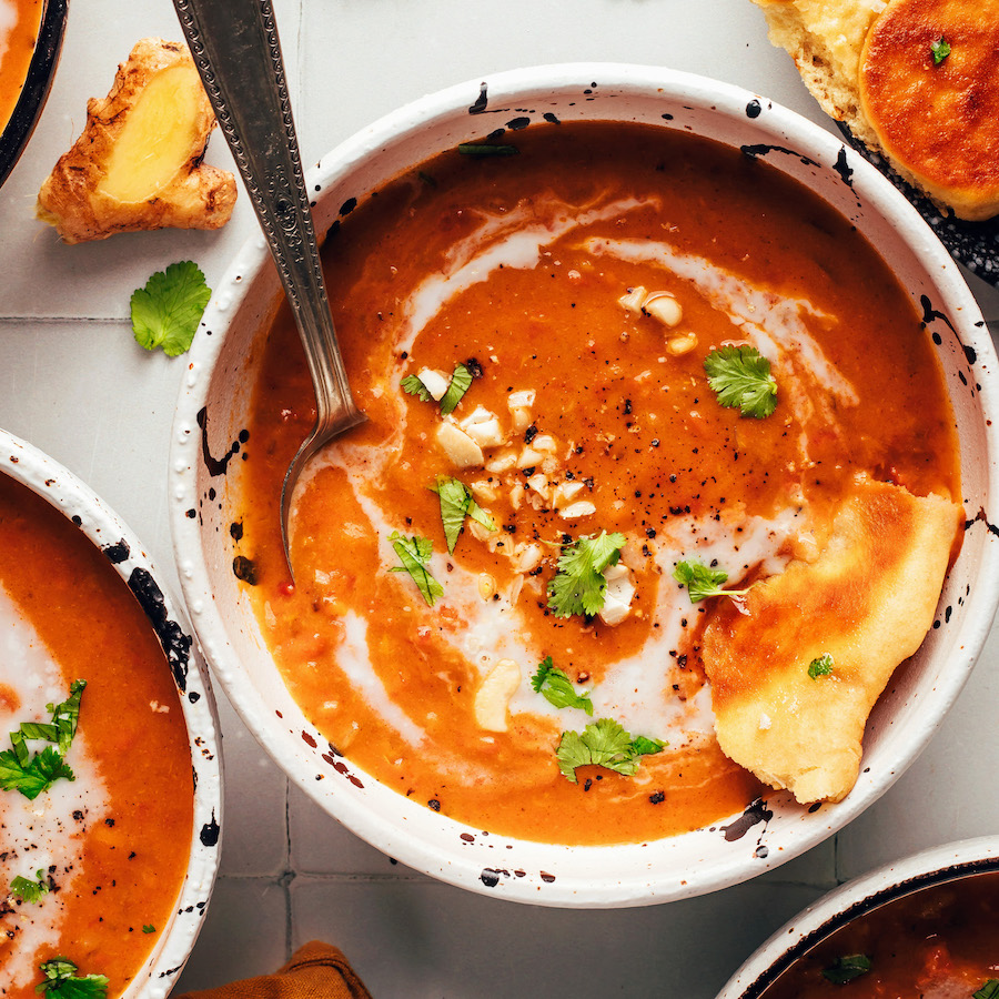 Overhead photo of a bowl of spicy pumpkin tomato soup with a swirl of coconut milk, toasted cashews, cilantro, and naan