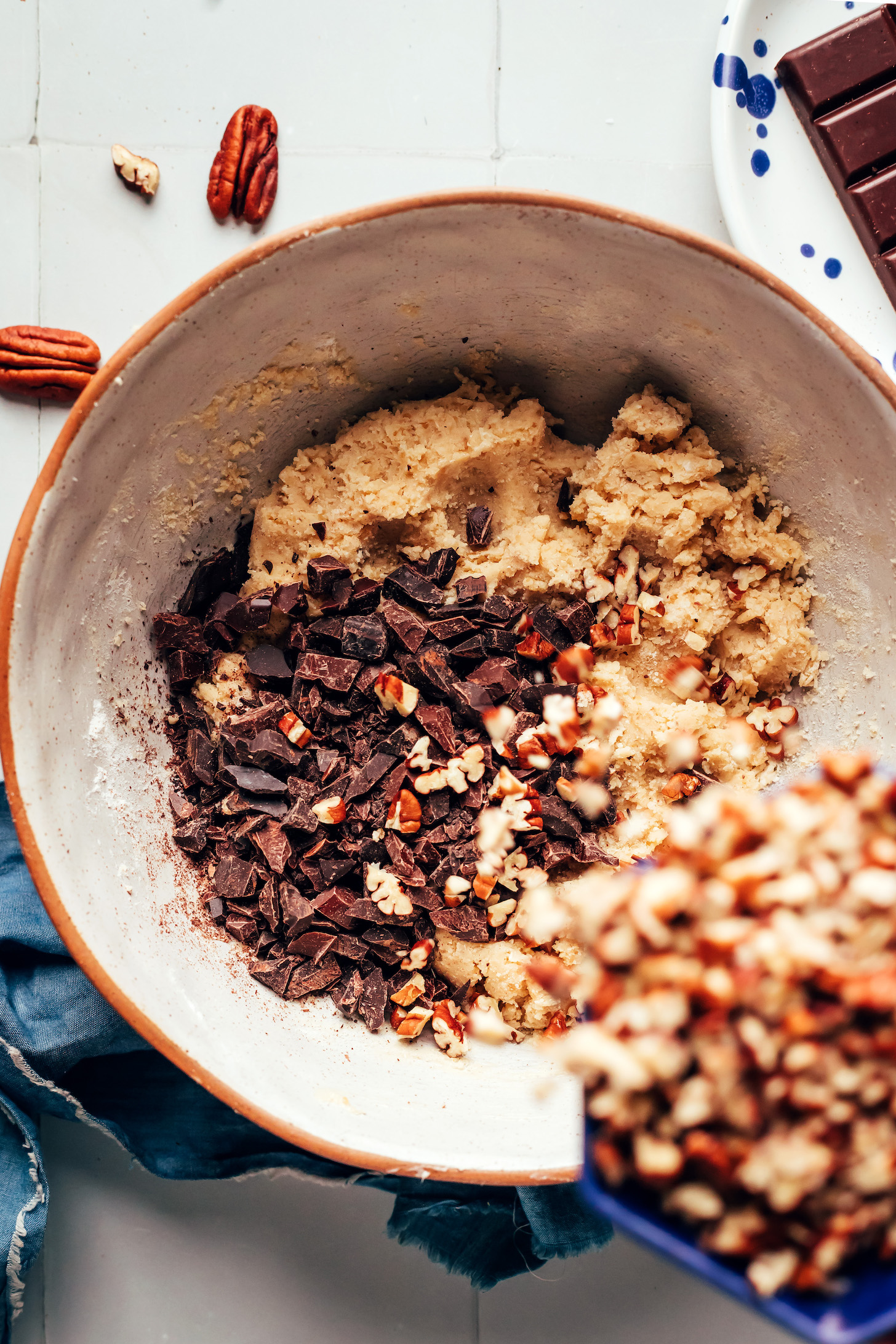 Pouring chopped pecans into a bowl of cookie dough topped with chopped dark chocolate