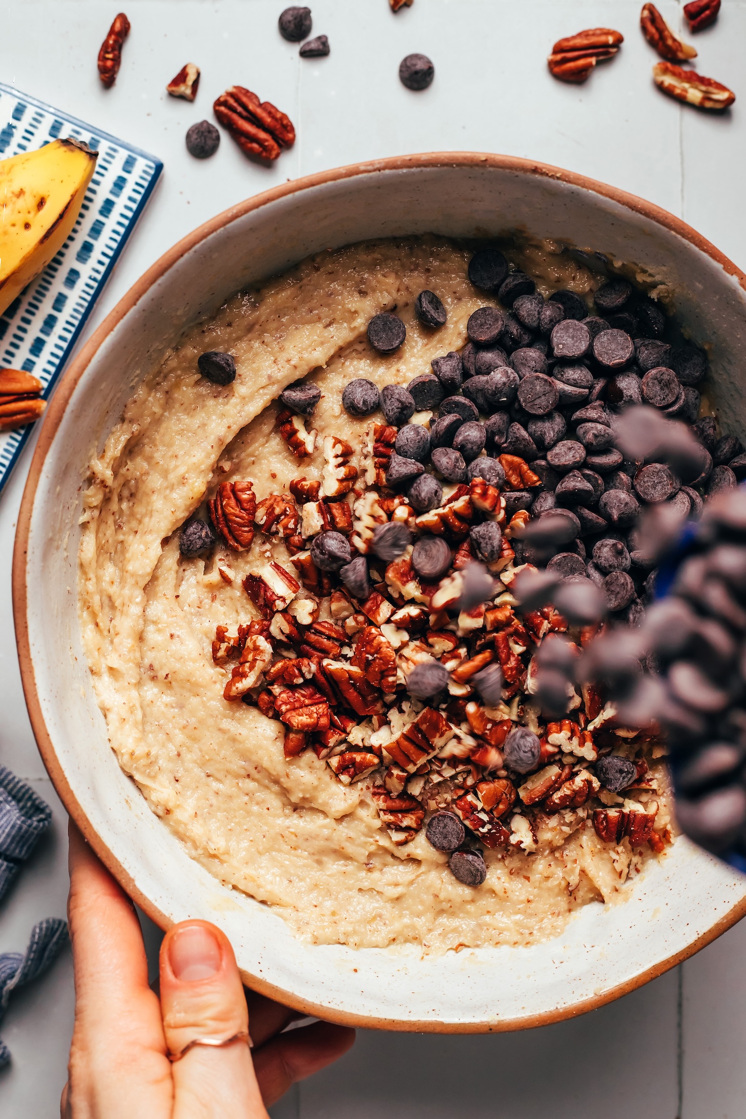 Adding chocolate chips into a bowl of batter topped with chopped pecans