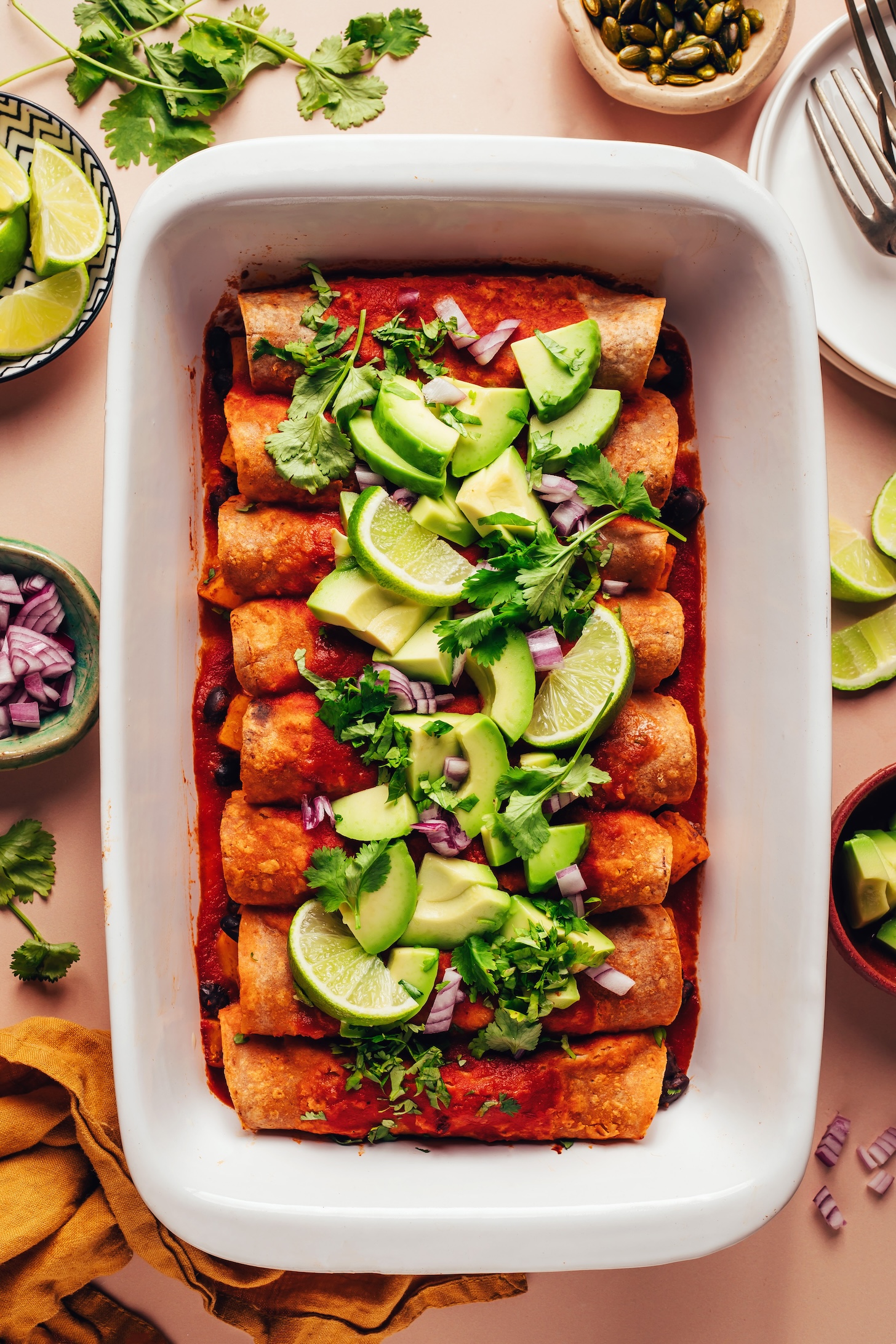 White ceramic casserole dish filled with butternut squash black bean enchiladas topped with avocado, cilantro, red onion, and lime