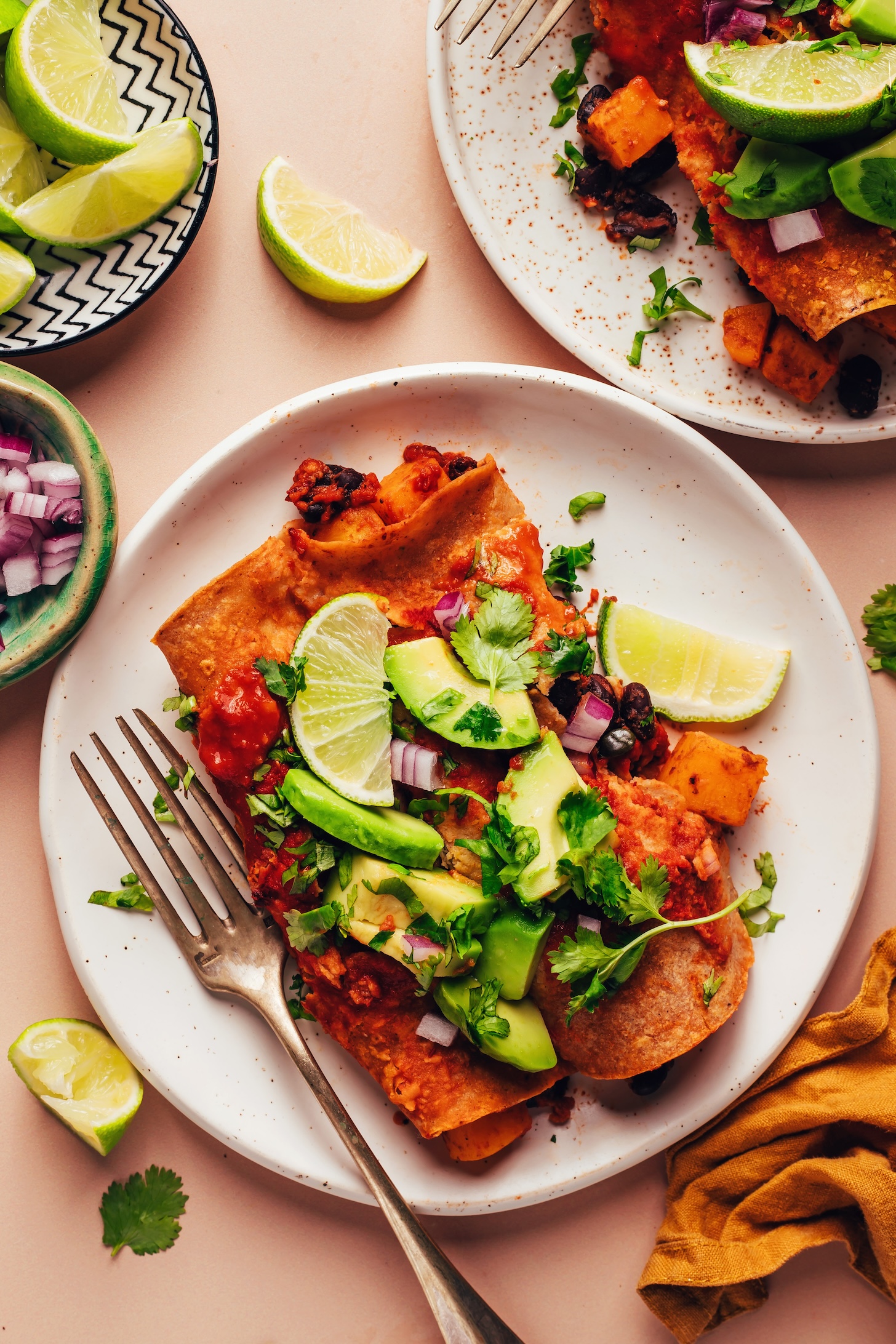 Two plates of butternut squash black bean enchiladas topped with avocado, red onion, cilantro, and lime wedges