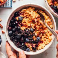 Hands holding the side of a bowl and a spoon in a bowl of creamy pumpkin oats with blueberries and almonds