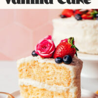 Slice of gluten-free dairy-free vanilla cake on a plate with more cake behind it on a cake stand