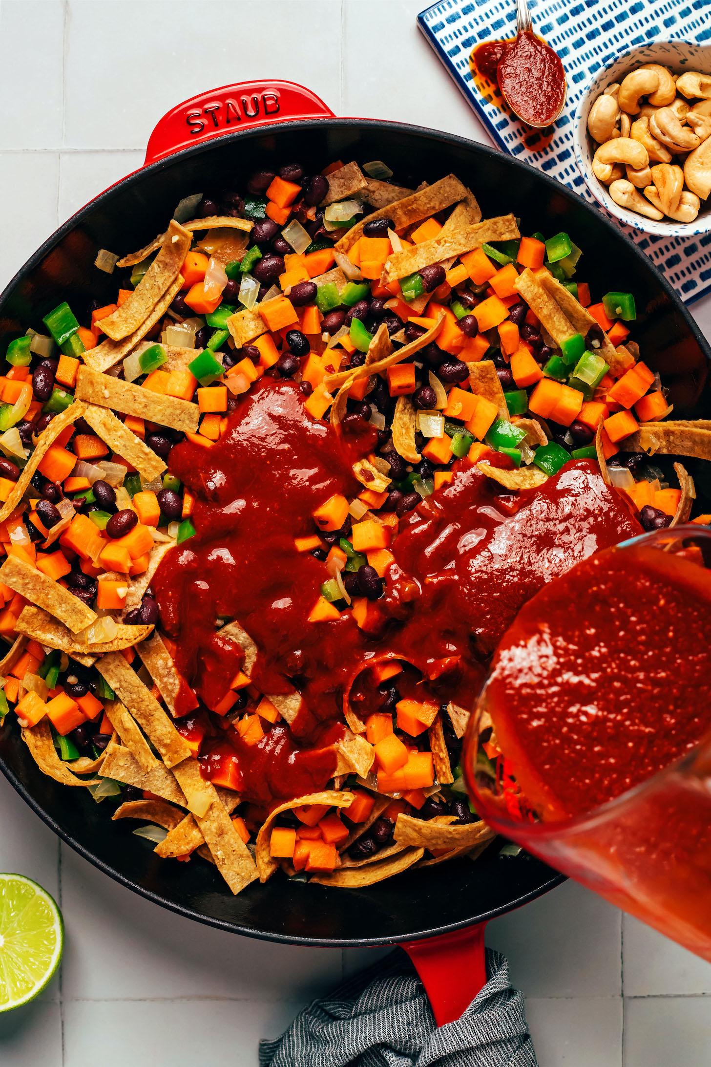 Pouring enchilada sauce over a skillet of sautéed vegetables, corn tortillas, and black beans