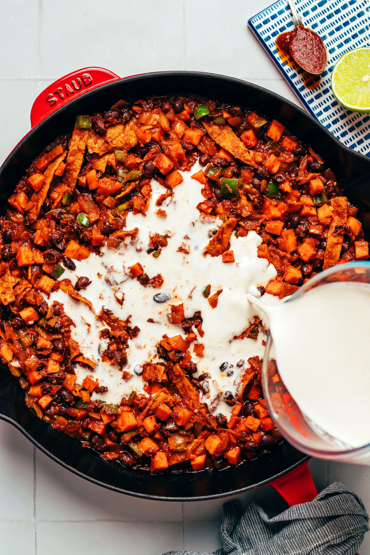 Pouring a creamy cashew sauce over a skillet of saucy sautéed vegetables, beans, and tortillas