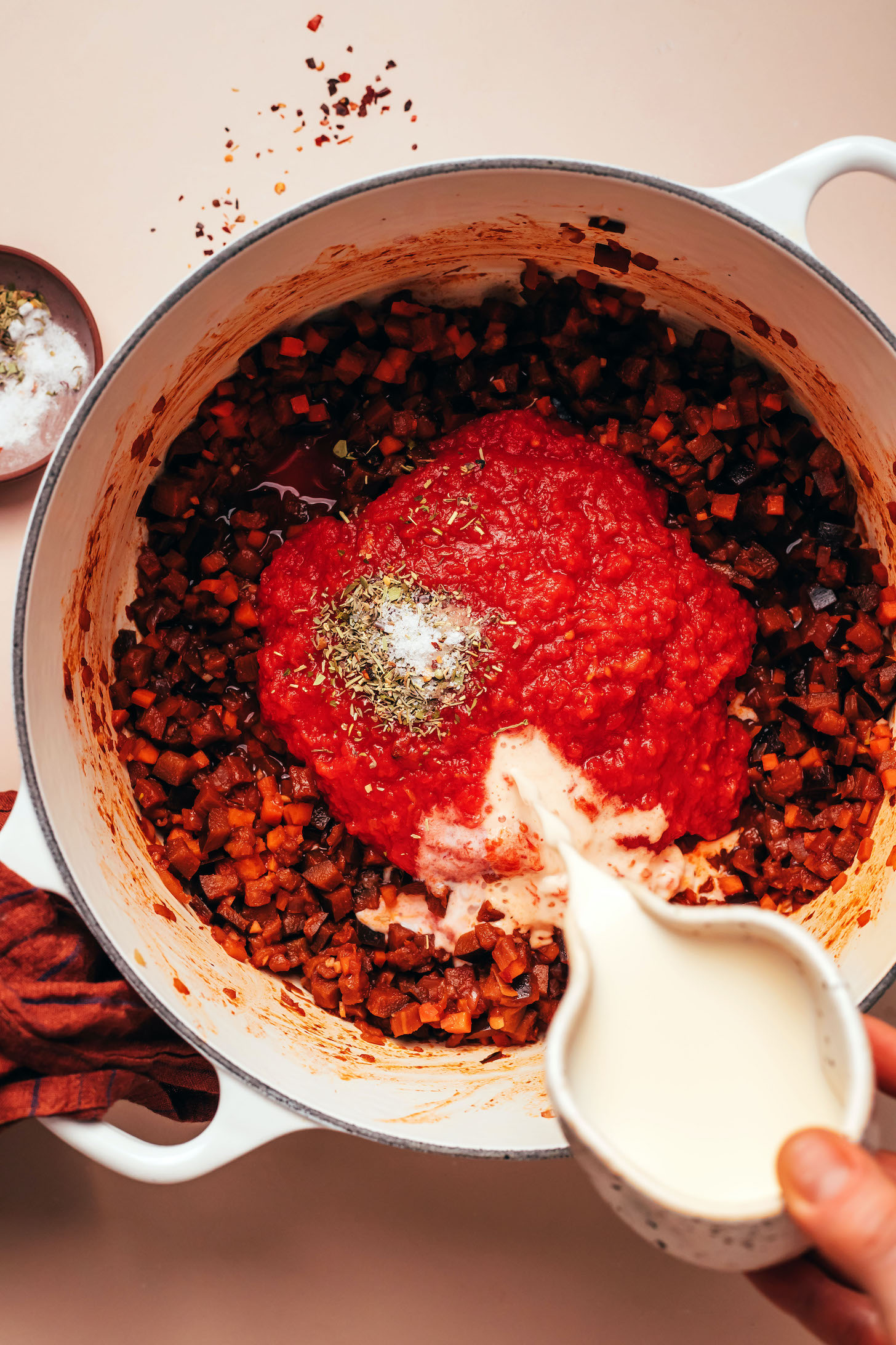 Pouring homemade cashew milk into a pot of crushed tomatoes, herbs, salt, and sautéed vegetables