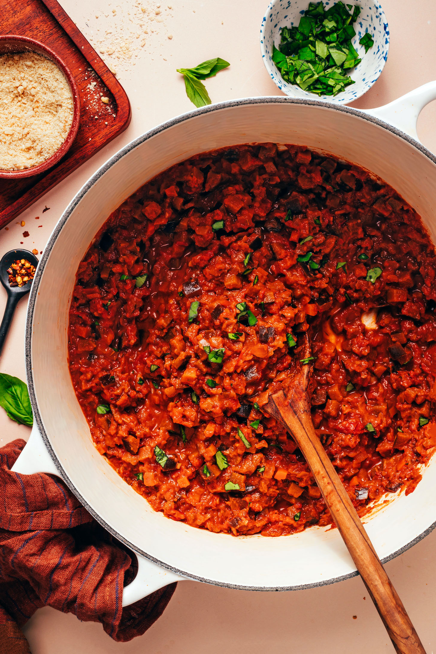 Batch of our vegetarian eggplant bolognese in a Dutch oven with red pepper flakes, vegan parmesan, and fresh basil around it